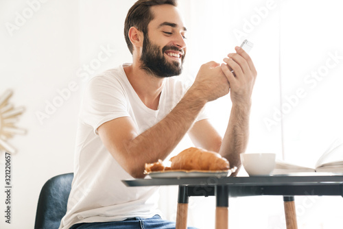 Man indoors at home have a breakfast using mobile phone.