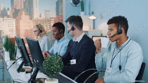 Customer support agent drinks tea and is bored. His multiracial colleagues answers the client's questions about the contract. They communicating with clients on computer or on phone.