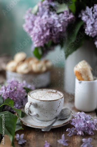 Cup of cappuccino and cake cones from puff pastry with vanilla cream in a metal box in spring still life with a bouquet of lilacs on a wooden table