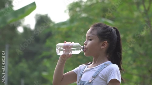 A little girl drinking clean water from a bottle against the background of trees. A happy little girl in a white t-shirt with a long haircut is smiling at the camera on the nature outdoors.