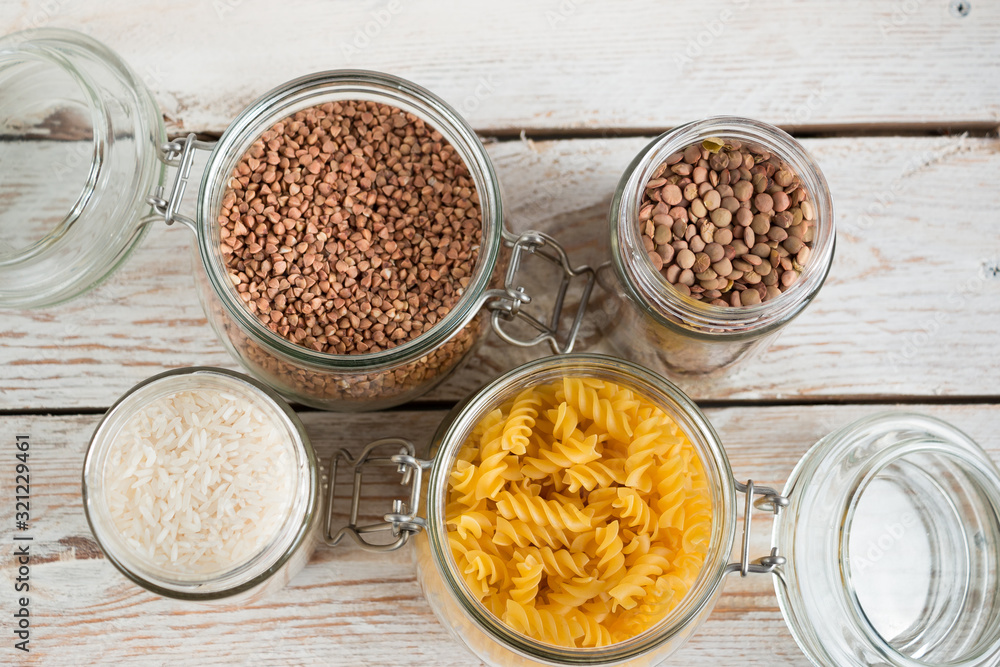 A variety of crops (buckwheat, rice, pasta, chickpeas) in glass jars on a wooden table on a light background. Close-up. The concept of vegetarianism and a balanced diet. A variety of crops (buckwheat, rice, pasta, chickpeas) in glass jars on a wooden table on a light background. Close-up. The concept of vegetarianism and a balanced diet.