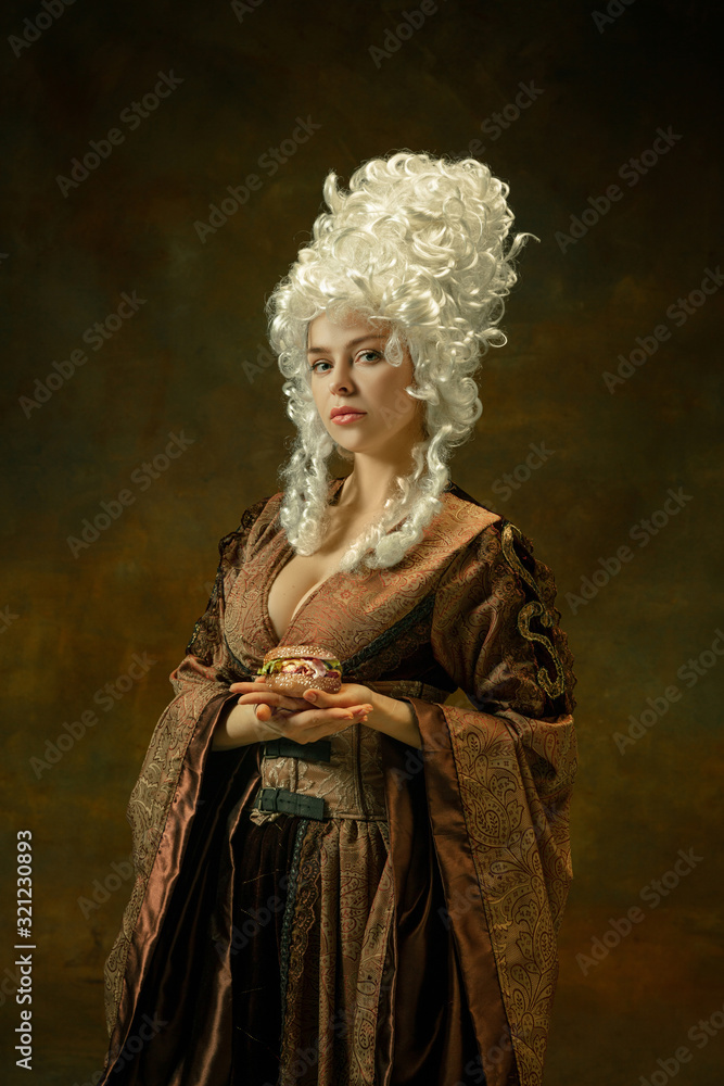 Calm, holding burger. Portrait of medieval young woman in brown vintage clothing on dark background. Female model as a duchess, royal person. Concept of comparison of eras, modern, fashion, beauty.