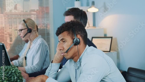 Bored multiethnic customer support operator waiting for telephone call from client. His colleagues are busy working.