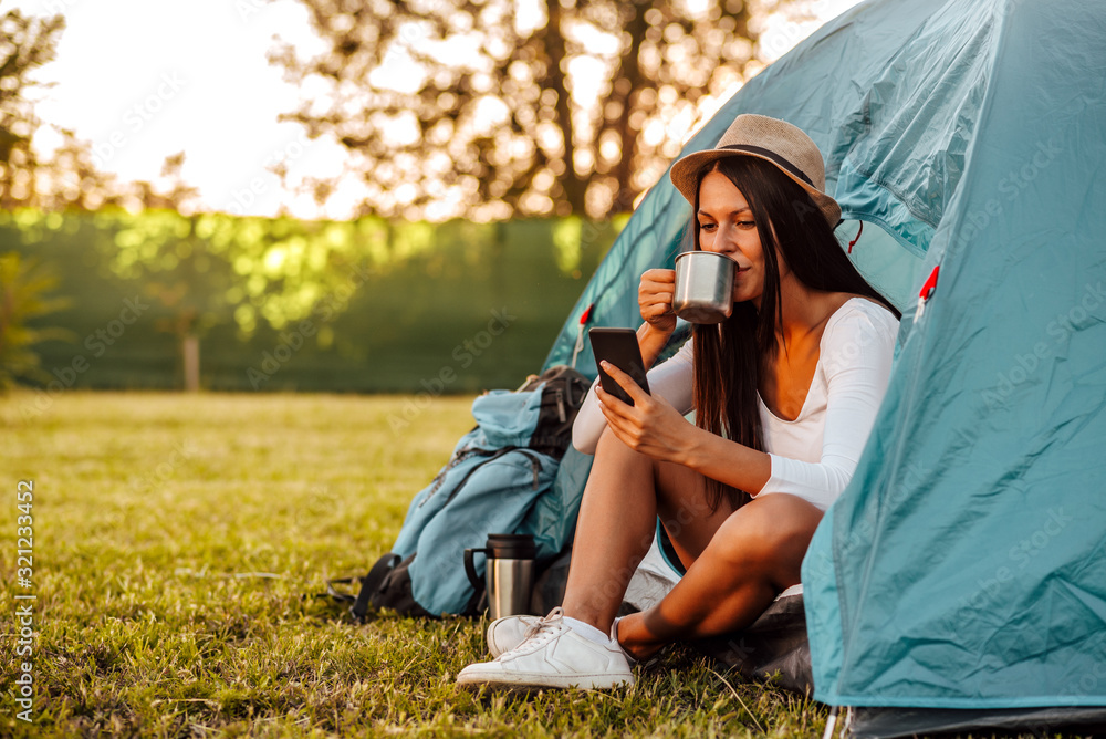 Portrait of a young woman at camping, sitting at tent drinking coffee and looking at smart phone. - obrazy, fototapety, plakaty 