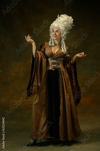 Elegance posing. Portrait of medieval young woman in brown vintage clothing on dark background. Female model as a duchess, royal person. Concept of comparison of eras, modern, fashion, beauty.