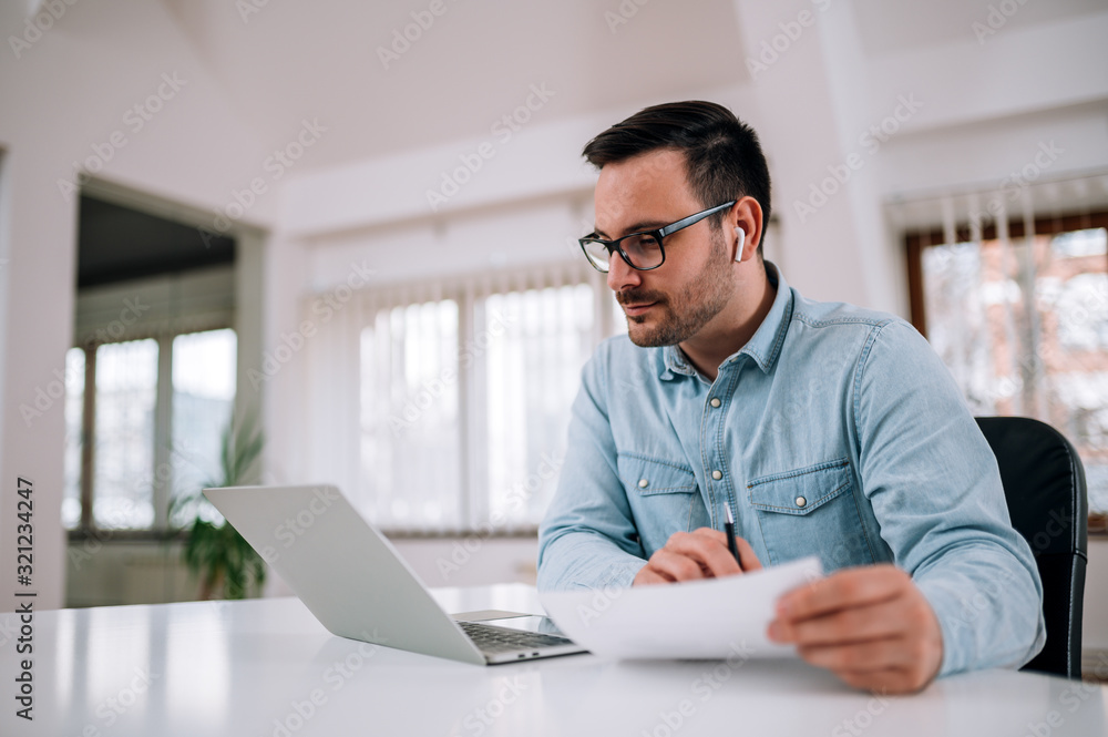 Entrepreneur looking at laptop and holding paper document in bright office, portrait. Entrepreneur looking at laptop and holding paper document in bright office, portrait.