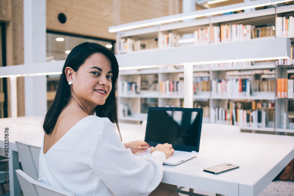 Portrait of prosperous female manager sitting in library using laptop computer for typing on job, smiling prosperous businesswoman looking at camera using modern technologies for research and work Portrait of prosperous female manager sitting in library using laptop computer for typing on job, smiling prosperous businesswoman looking at camera using modern technologies for research and work