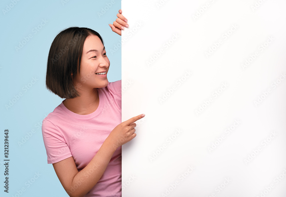 Cheerful Asian woman pointing at blank poster Cheerful Asian woman pointing at blank poster