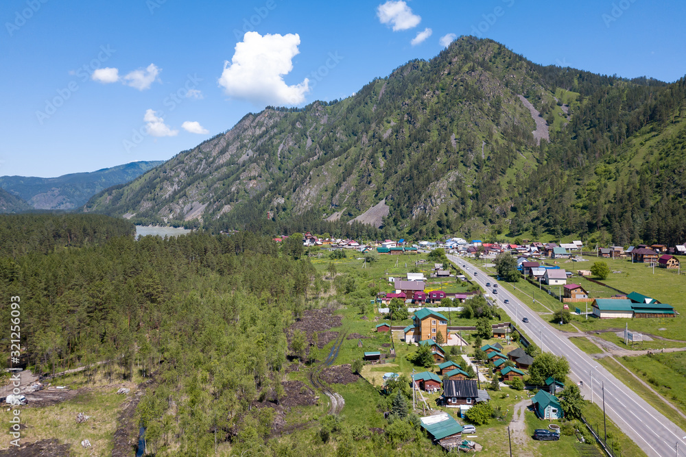 Aerial view of country houses in the village near the highway and the river against the backdrop of the mountains under the blue sky on a warm summer summer day. Picturesque landscape on vacation.