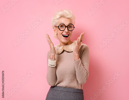 Happy grandmother raises palms from joy and shouts loudly. Photo of kind elderly woman wears eyeglasses on pink background.