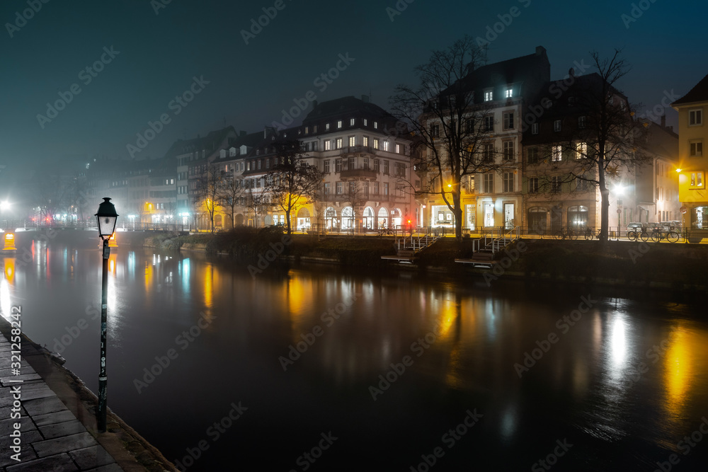 Obraz premium Ile River Embankment in Strasbourg at night, fog. Reflections of illuminated buildings and lanterns in the water
