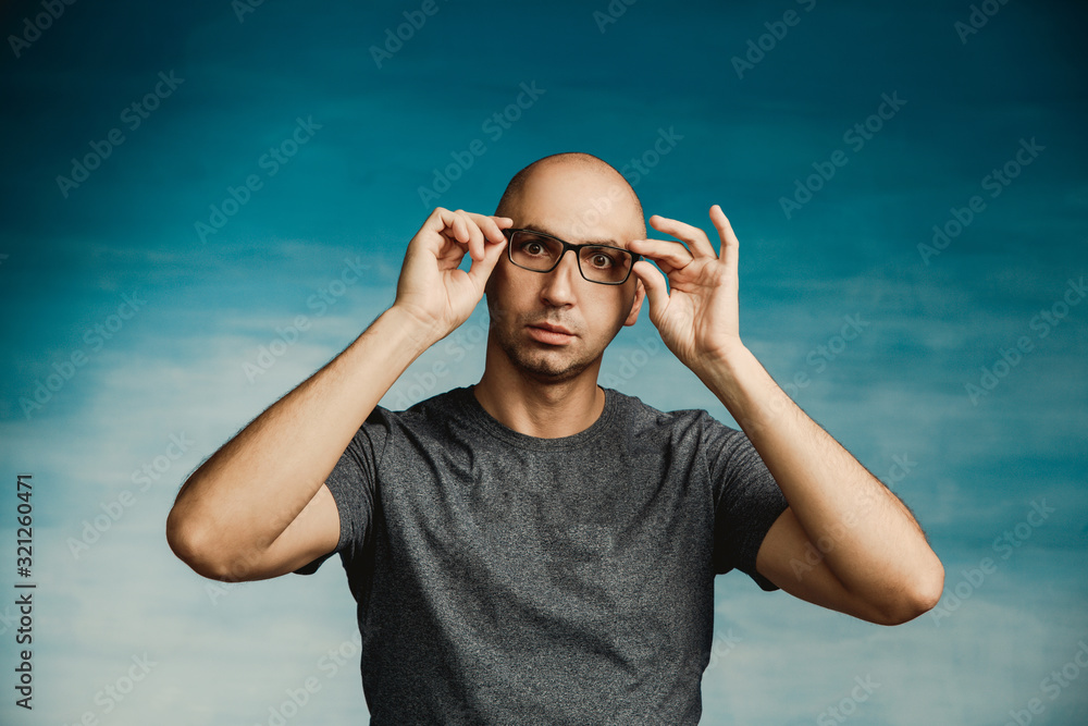 Naklejka premium An interesting bald man adjusts his glasses with his hands and looks in surprise at the camera on a blue background.