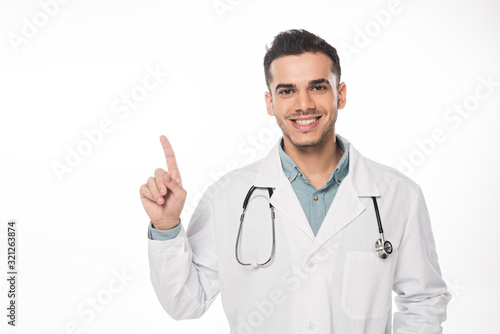 Handsome doctor pointing with finger and smiling at camera isolated on white
