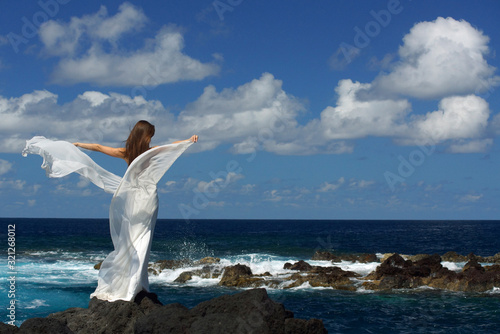 Back view of young bride with white wings of wedding dress on rock sea shore. Ocean waves, splash of water