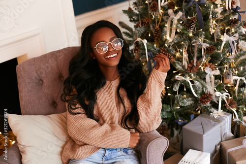 Happy fashionable black girl in a luxurious Christmas interior. African American woman sitting in a stylish armchair against the background of the Christmas tree.