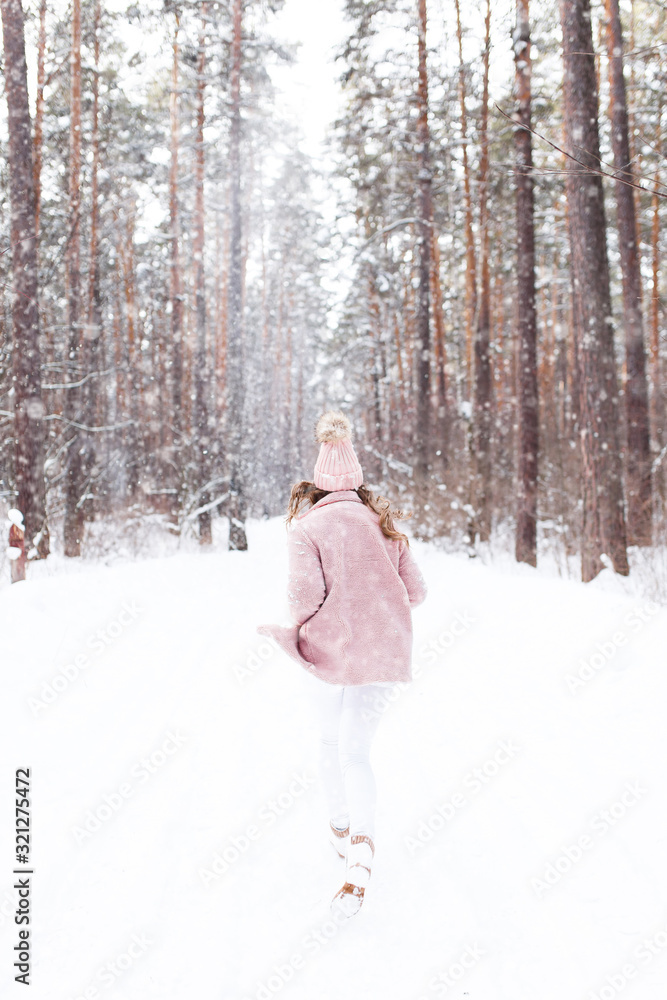A beautiful pretty young girl with curly hair and a snow-white smile in a pink fur coat is walking and fooling around in the winter forest against the background of snow and trees, enjoy