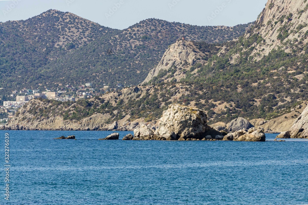 Panorama from city embankment on rock in sea near village of New World. Birds are sitting on rock. Urban-type settlement against backdrop of Crimean mountains. Sudak, Crimea, Russia, October , 2019.
