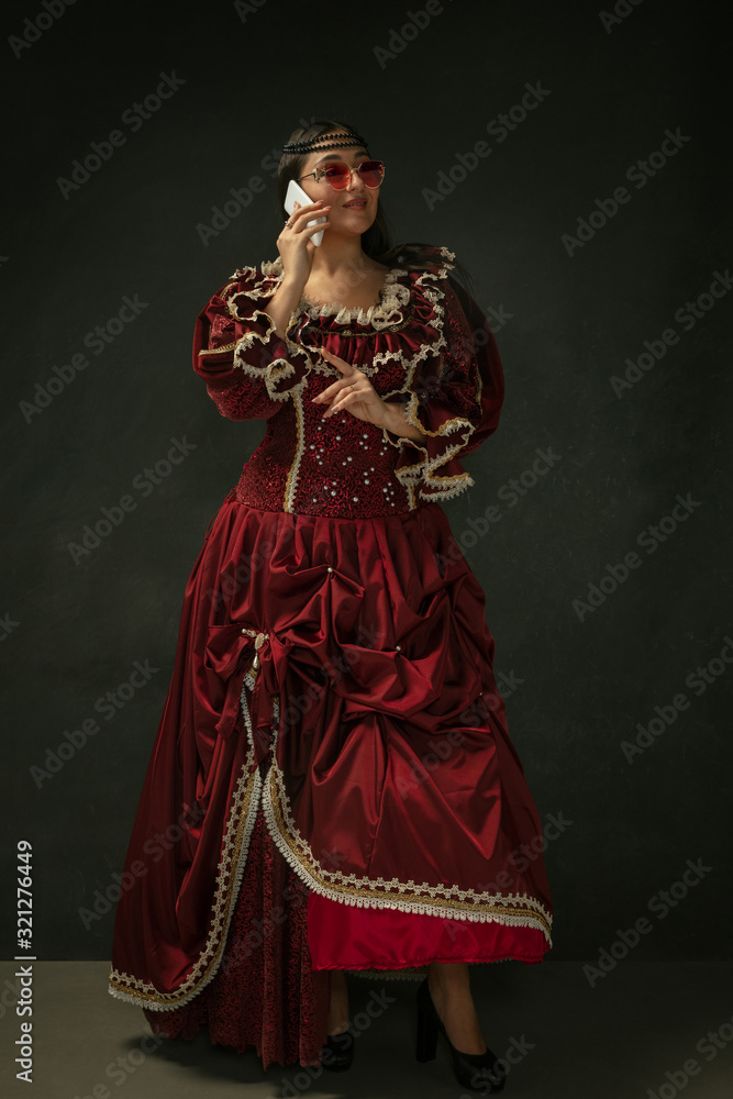 Talks on phone wearing modern eyewear. Medieval young woman in red vintage clothing on dark background. Female model as a duchess, royal person. Concept of comparison of eras, modern, fashion, beauty.