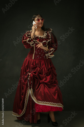 Talks on phone wearing modern eyewear. Medieval young woman in red vintage clothing on dark background. Female model as a duchess, royal person. Concept of comparison of eras, modern, fashion, beauty.