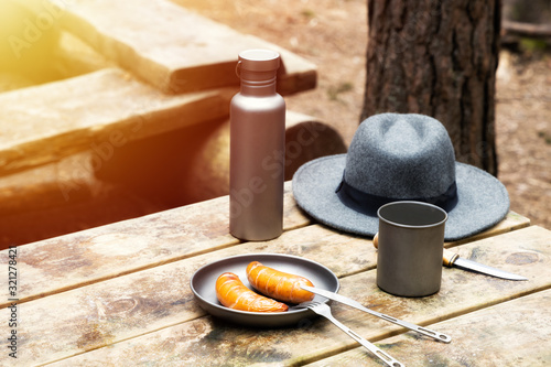 Knife, hat, water bottle and titanium mug on the wooden table. Grilled sausages on the plate. Top view. Outdoors. Bushcraft concept.