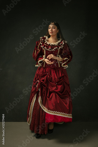 Posing as royal. Portrait of medieval young woman in red vintage clothing standing on dark background. Female model as a duchess, royal person. Concept of comparison of eras, modern, fashion, beauty.