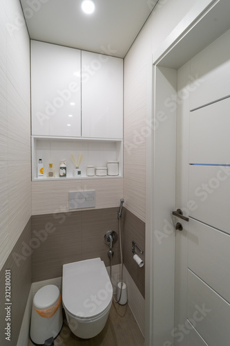 Modern design toilet with beige and brown wall and floor tiles, wooden doors, white built-in toilet bowl, a bin, bidet shower, scrub brush and lamp in the ceiling. There are white wall cabinet and she