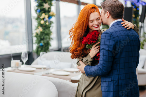 kind and gallant caucasian man in tuxedo invited on a dtae and gave red flowers, roses to his lady in dress. smiling pleasant redhaired woman thanks him. in restaurant