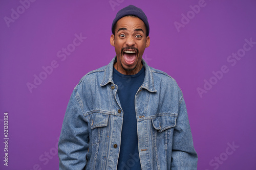 Furious young brown-eyed bearded man with dark skin shouting angrily and wrinkling forehead while looking heatedly at camera, wearing blue cap, pullover and jeans coat over purple background