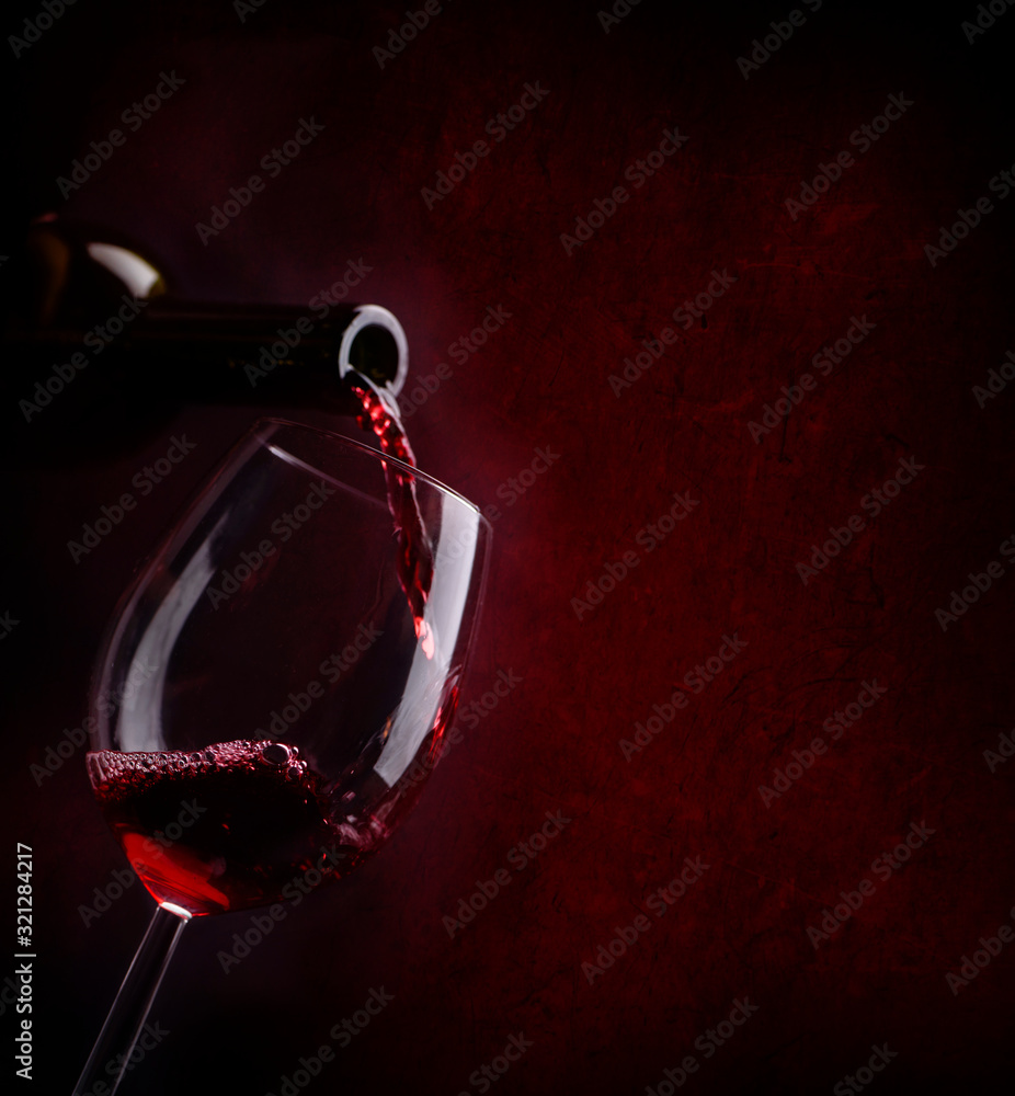 Fototapeta premium Red wine pouring in wineglass from bottle over dark background.