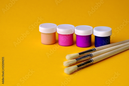 Colorful vibrant cans of gouache paint and brushes isolated on color yellow background. Colorful paint cans set, Painting background. A set of acrylic paints in jars for the artist. Copy space.