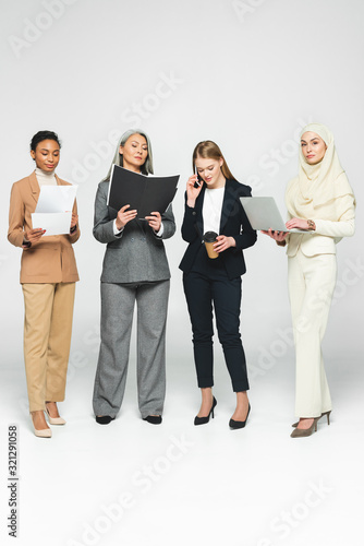 attractive multicultural businesswomen with folders near woman talking on smartphone on white