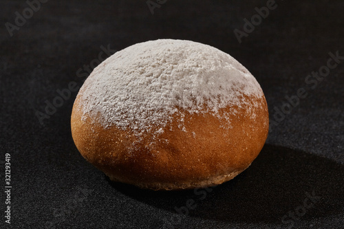 Fresh, tasty baked bun sprinkled with flour or powdered sugar against black background with copy space. Rural cuisine or bakery. Close-up