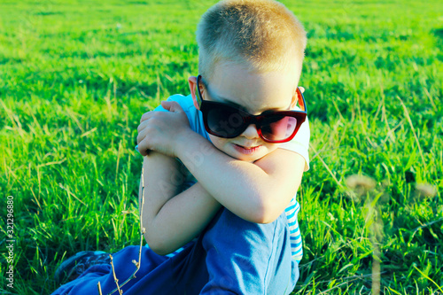 Unhappy little boy wearing sunglasses sitting on green grass. Cute little boy outdoors. People, emotions concept.