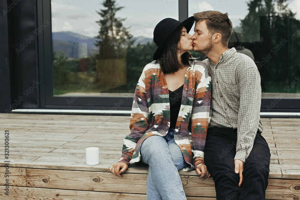 Stylish hipster couple kissing, sitting on wooden porch, relaxing on background of modern cabin with big windows in mountains. Happy young family travelers enjoying vacation in woods