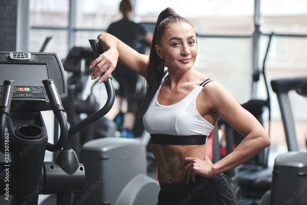 Strong sexy brunette woman with fitness muscular body posing near equipment in gym, concept of sports nutrition and supplements Strong sexy brunette woman with fitness muscular body posing near equipment in gym, concept of sports nutrition and supplements