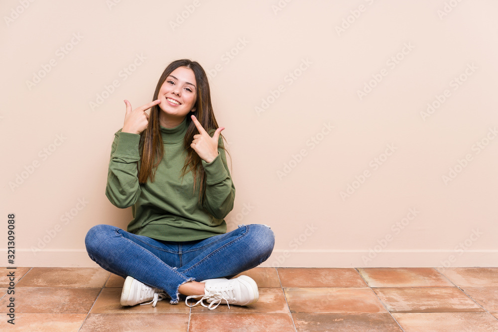 Young caucasian woman sitting on the floor isolated smiles, pointing fingers at mouth. Young caucasian woman sitting on the floor isolated smiles, pointing fingers at mouth.