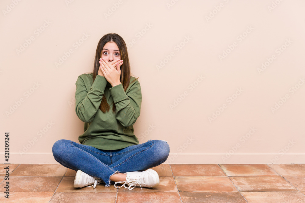 Young caucasian woman sitting on the floor isolated shocked covering mouth with hands. Young caucasian woman sitting on the floor isolated shocked covering mouth with hands.