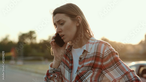 Disappointed young woman talking on the phone standing in the street at road accident. Mixed-up girl in shirt asking for help calling for the police. Car crash.