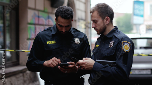 Police at raid. Close-up two handsome policemen searching on smartphones tracking a criminal online. Friendly colleagues cops at police investigation area in the city street.