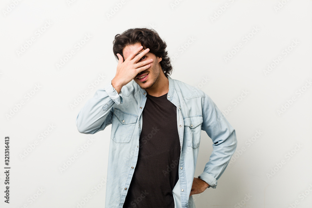 Young handsome man against a white background blink at the camera through fingers, embarrassed covering face. Young handsome man against a white background blink at the camera through fingers, embarrassed covering face.
