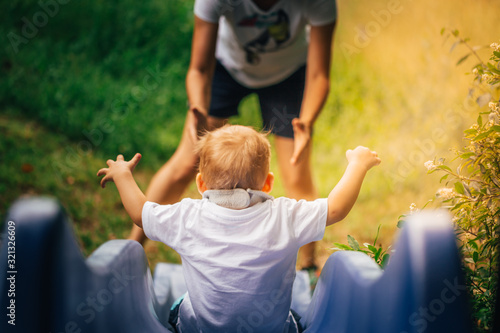 Little child sliding down a blue slide into the arms of the mother on a summer day. View from behind.