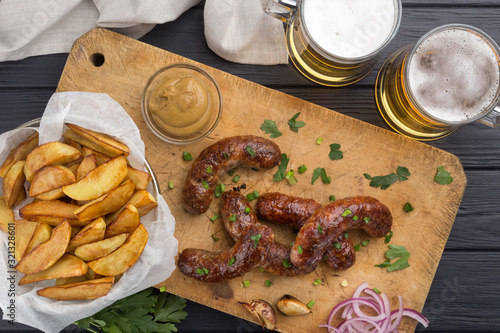 Grilled sausages from a BBQ served with a mustard, fried potatoes, garlic, herbs, beer on a wooden board all served on a black wooden table. Delicious german sausages Perfect for Octoberfest.