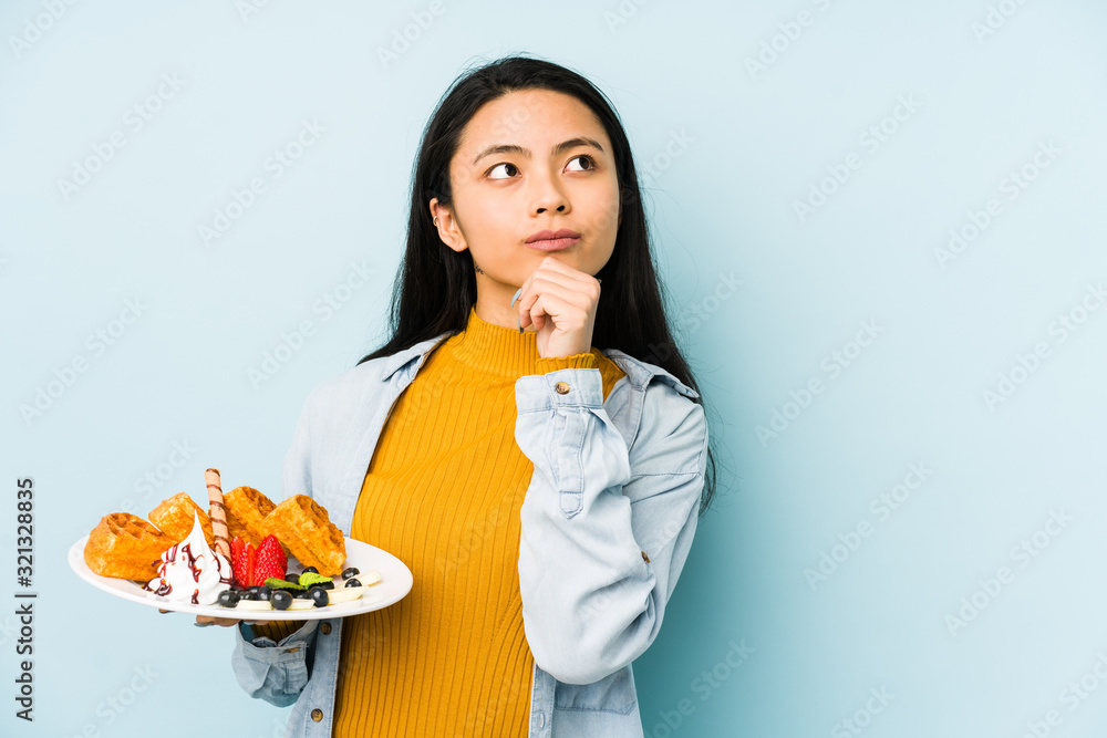Young chinese woman holding waffle isolated on blue background showing number one with finger. Young chinese woman holding waffle isolated on blue background showing number one with finger.