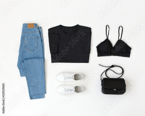 Blue jeans, black t-shirt, lace bra, cross body bag and sneakers on white background. Overhead view of woman's casual outfit. Trendy simple basic minimalistic look. Women clothes. Flat lay, top view.