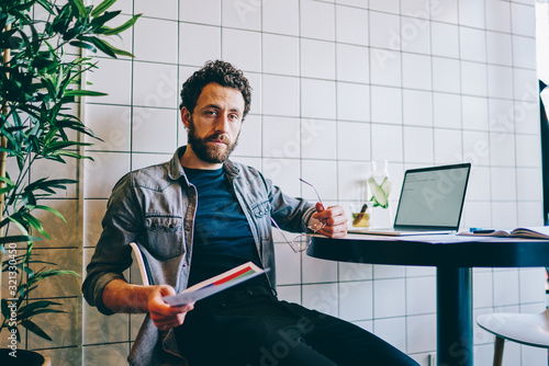 Tired man sitting at table with laptop and holding paper document