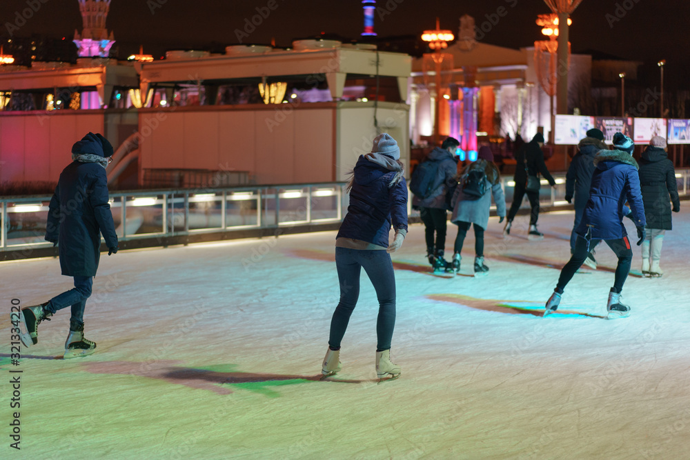 Fototapeta premium Ice skating rink in the city park. Men and women are here. Youth skating and getting fun in night time. Sportive lifestyles concepts. Backs.