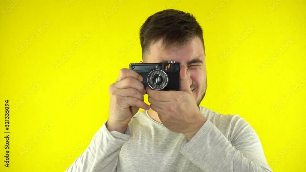 A young man smiles and takes a picture using a vintage camera. Handsome caucasian male in t-shirt taking photo with old retro film camera. Yellow plain background. A young man smiles and takes a picture using a vintage camera. Handsome caucasian male in t-shirt taking photo with old retro film camera. Yellow plain background.