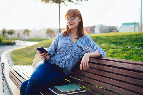 Charming woman in casual wear using smartphone while relaxing on bench at park
