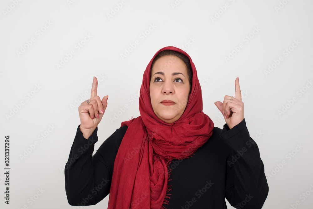 Horizontal shot of successful friendly looking young dark haired woman with bright make up exclaiming excitedly, pointing both index fingers up, indicating something shocking on blank copy space wall. Horizontal shot of successful friendly looking young dark haired woman with bright make up exclaiming excitedly, pointing both index fingers up, indicating something shocking on blank copy space wall.
