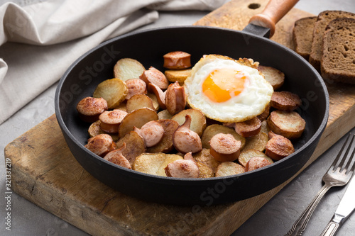 Close-up shot of fried potatoes with sausages and egg. Breakfast. Potatoes with sausages, cut into slices and fried in oil, in rustic style. Frying pan with tasty cooked egg and sausages on table